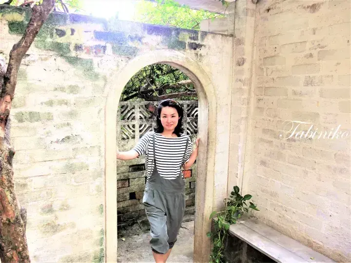 A woman in striped shirt and overalls stands in an arched doorway of a rustic brick building with greenery.