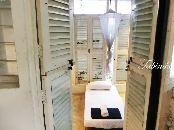 A serene massage room with a white treatment bed, mosquito net, and white louvered shutters at Tabiniko.