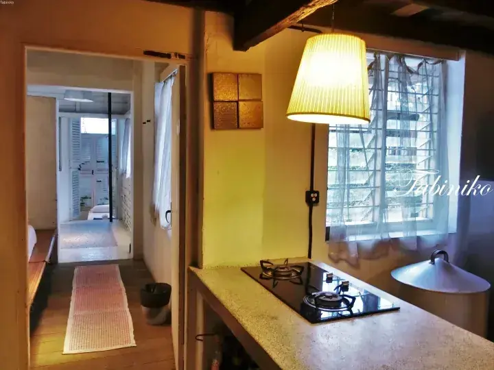 Cozy kitchenette with a gas stove and hanging lamp, featuring a view into a hallway with wooden flooring and a window with sheer curtains.