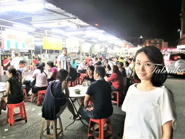 A woman smiles at a bustling night food market in Malaysia, surrounded by diners at red plastic stools under bright lights.