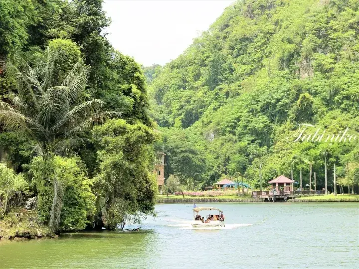 A boat tour glides across the calm waters of Tasik Puteri in Ipoh, surrounded by lush green hills and tropical vegetation.