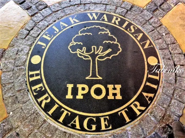 舗装された地面に埋め込まれた「Jejak Warisan Ipoh Heritage Trail」のマーキング。木と金色の文字が特徴。