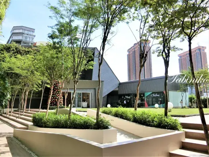 Modern building exterior with landscaped garden, trees, and steps, with high-rise towers in the background.