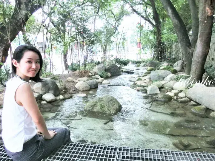 Woman sitting on a metal grate beside a clear stream with rocks and trees in a lush park setting.