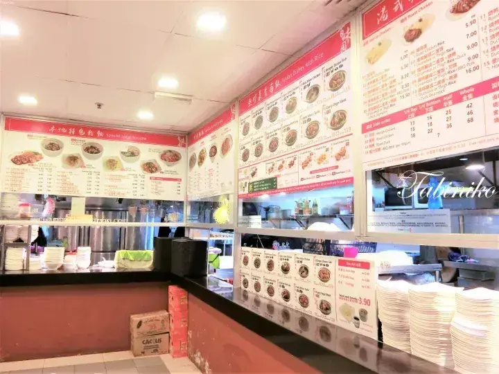 Interior view of a Malaysian hawker centre food stall with red menu boards displaying dishes and prices above the serving counter.