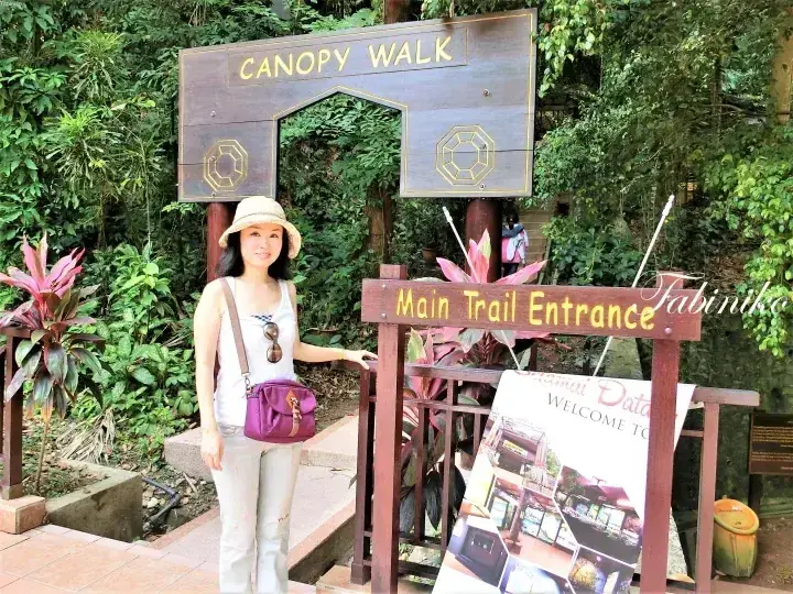 A woman stands at the Main Trail Entrance sign for the Canopy Walk in a lush tropical forest.