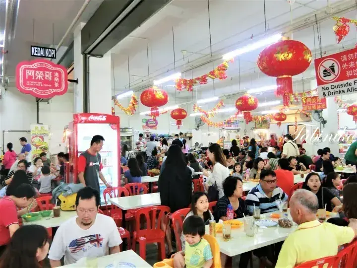 Crowded hawker centre with diners eating at tables under festive red lanterns and a KEDAI KOPI sign.