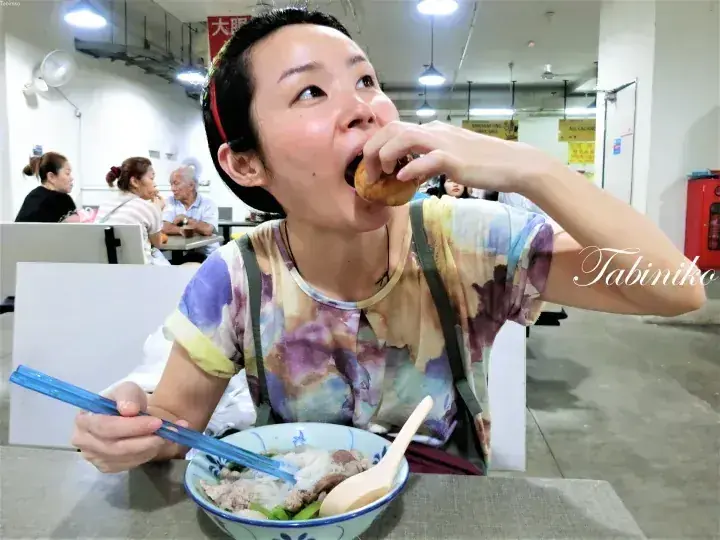 A woman eats a fried snack while seated at a table with a bowl of noodles in a local Malaysian food court.