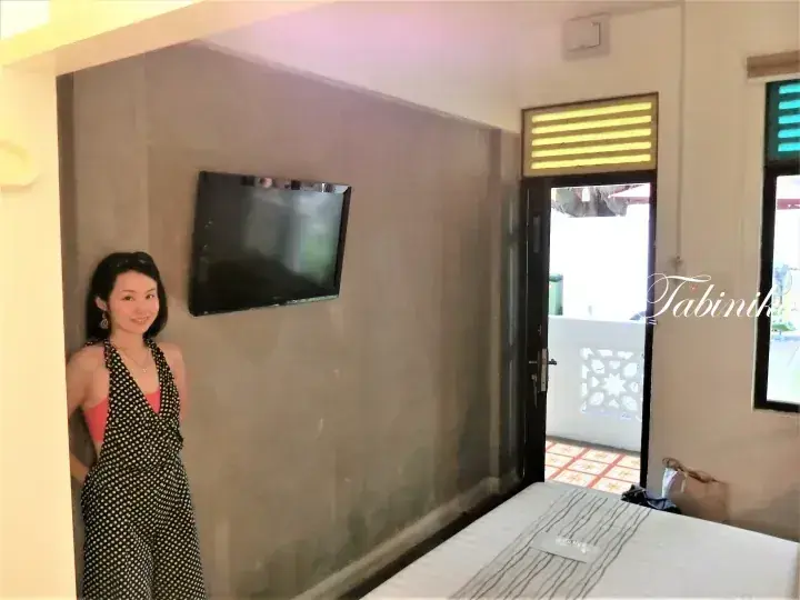A woman poses in a stylish jumpsuit inside a modern hotel room with a wall-mounted TV and balcony access.