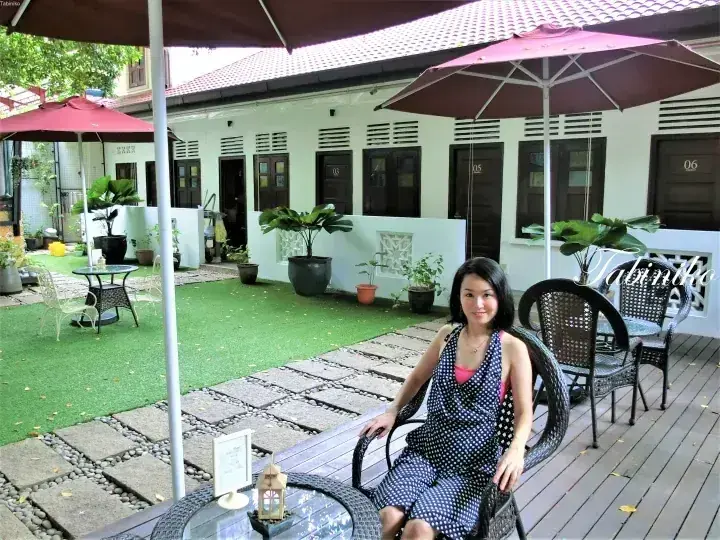 A woman sits on a wooden deck at a boutique hotel courtyard with green lawn, umbrellas, and numbered rooms.