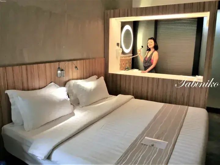 A woman stands in a lit vanity mirror niche above a neatly made double bed in a modern hotel room.