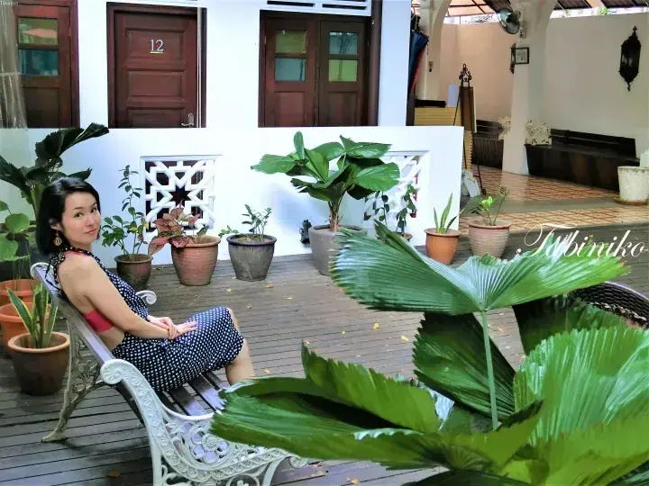 A woman relaxes on a white bench on a wooden deck surrounded by potted plants at a colonial-style property.