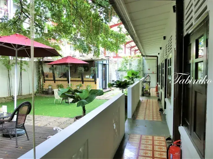 Outdoor corridor with tiled flooring and a fire extinguisher overlooking a courtyard with green turf, red umbrellas, and lush trees.