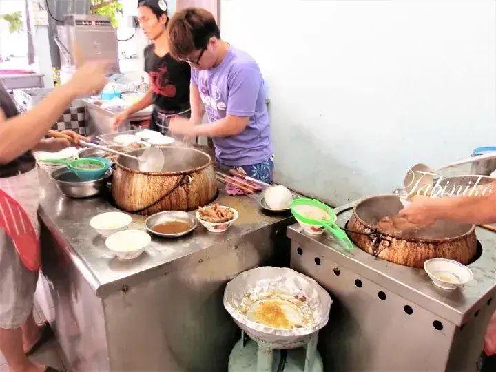 Street vendors preparing traditional Malaysian food at a busy hawker stall with large cooking pots.