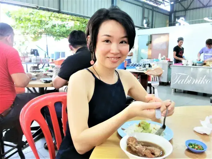 A smiling woman enjoys a meal of soup and rice at an outdoor eatery with a Tabiniko counter visible in the background.