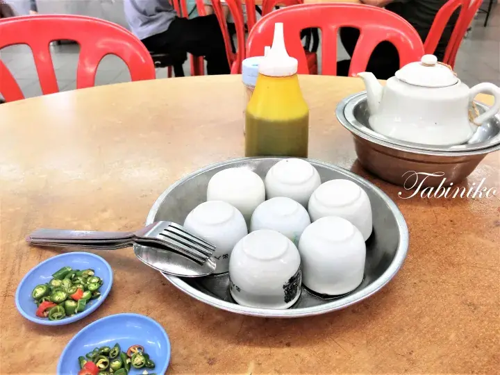 マレーシアのコピティアムで木製のテーブルに置かれた白い蒸しパン、フォーク、唐辛子ソース、茶壺。