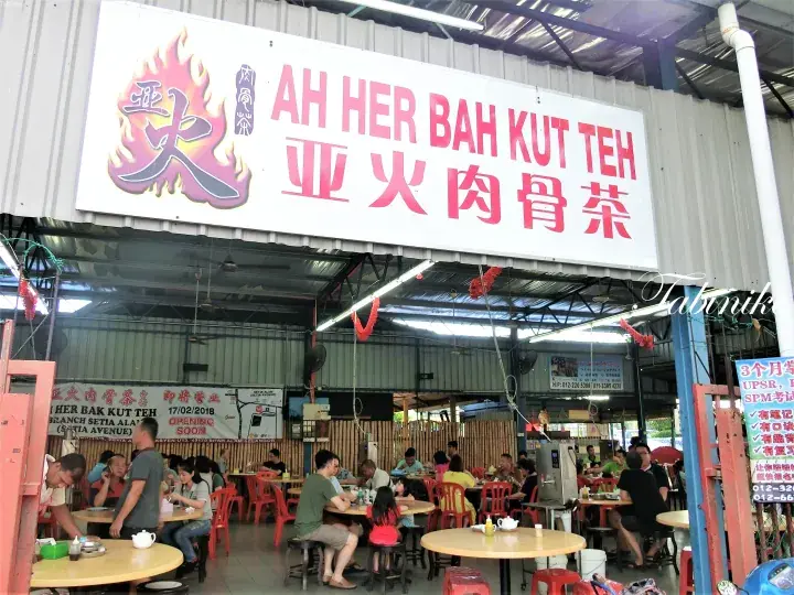 屋外席で食事をする客がいる「Ah Her Bah Kut Teh」の看板と店舗の外観。