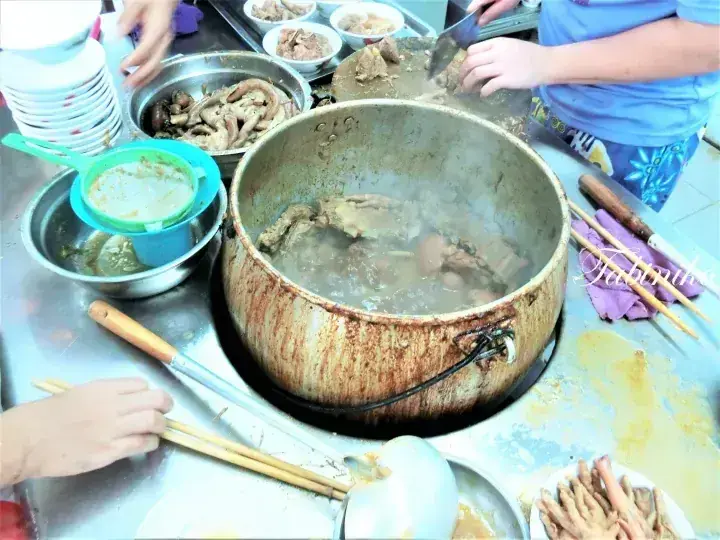 A large pot of simmering meat soup on a stove with bowls of ingredients nearby