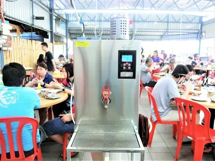 A stainless steel water dispenser stands in a busy Malaysian kopitiam with patrons dining at round tables.