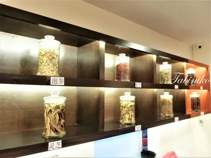 Glass jars containing dried herbs like Lingzhi and Paoshen displayed on dark wooden shelves in a traditional medicine shop.