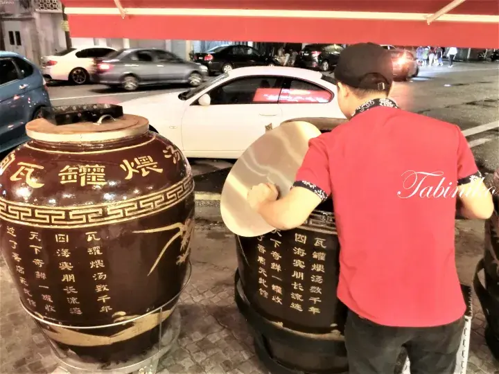 赤いシャツの男性が屋台で大きな木べらを使って伝統的な土鍋スープを混ぜている。