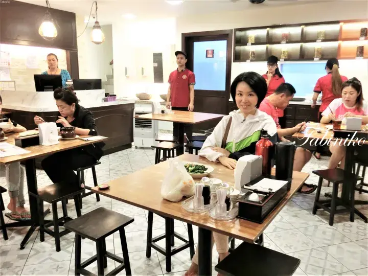 赤い制服を着たスタッフや他の客がいる賑やかなレストランで、テーブルに座る女性。