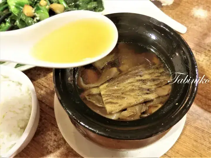 A spoon lifts golden broth from a claypot dish featuring braised fish and vegetables, served with rice.