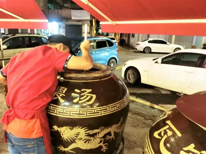赤いシャツの男性が夜店で大きな龍のデザインが描かれた陶器の鍋を混ぜている。
