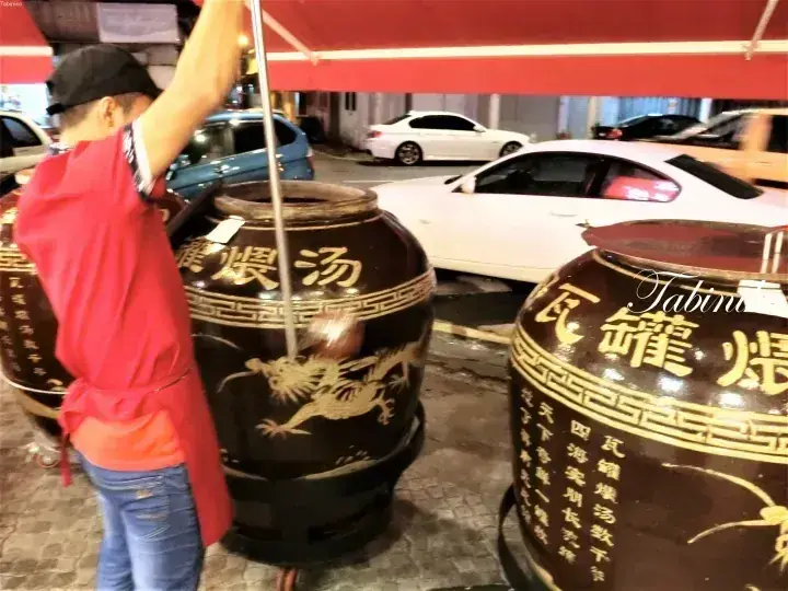 A vendor in a red shirt stirs a large ceramic claypot soup at a night market stall.