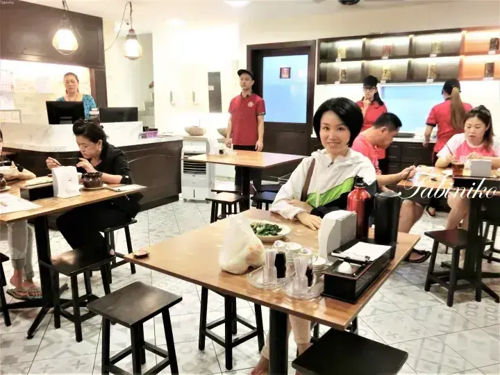 A woman smiles at a table in a busy restaurant with staff in red uniforms and other diners in the background.