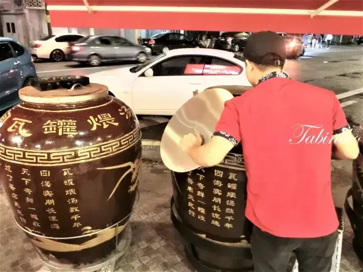 A man in a red shirt uses a large wooden paddle to stir a traditional clay pot soup at a street stall.