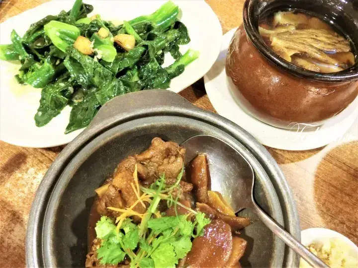 A traditional Malaysian meal featuring claypot chicken, stir-fried greens, and a claypot soup served on a wooden table.
