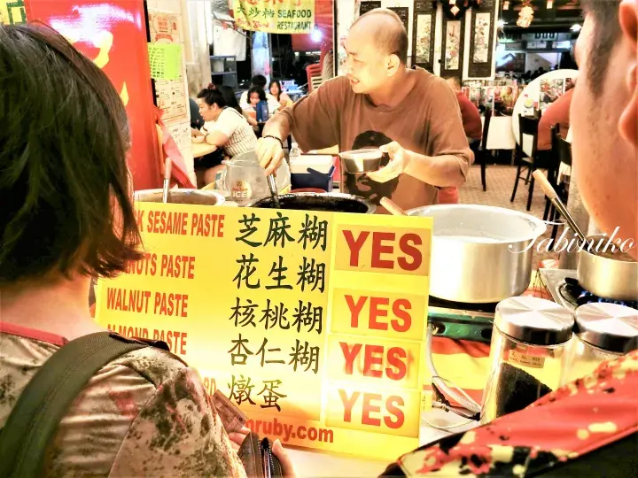 黒胡麻ペーストやクルミペーストなどの伝統的なマレーシアのデザートを黄色いメニュー看板の前で提供する屋台の店主。