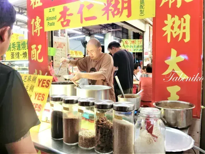 A vendor prepares almond paste at a traditional stall in a Malaysian hawker center, surrounded by jars of ingredients.