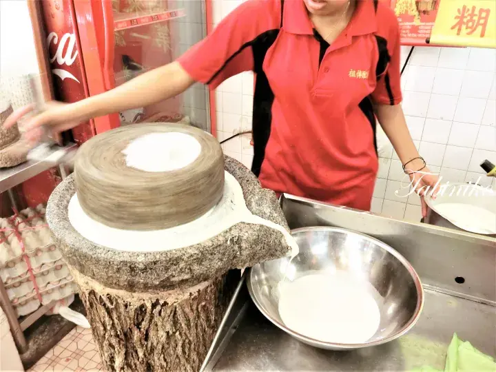 マレーシアの朝食屋台で、伝統的な石臼を使って新鮮な豆乳を挽く作業風景。