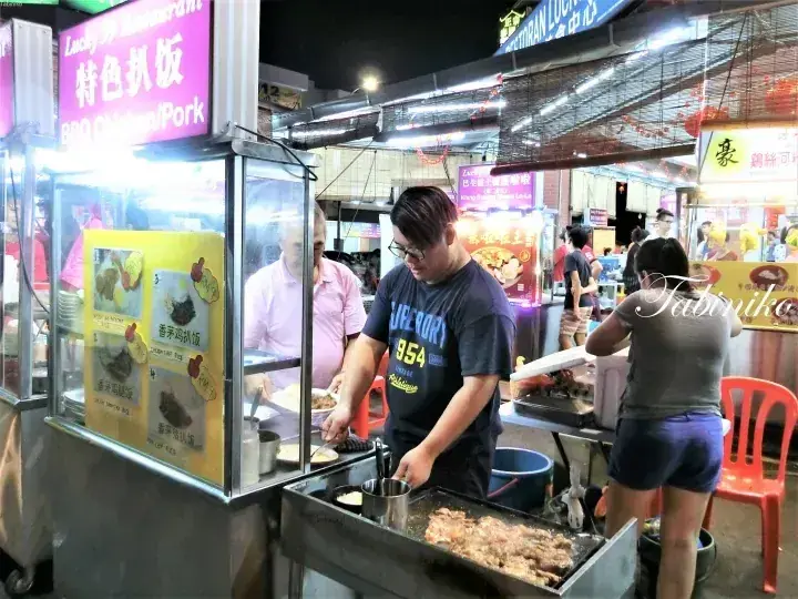 A vendor grills meat at a street food stall with a sign reading &ldquo;Lucky Restaurant BBQ Chicken/Pork&rdquo; in a night market.