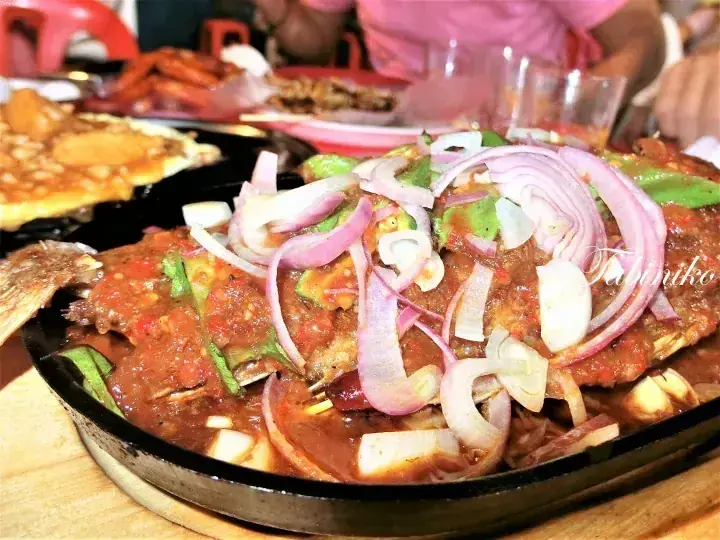 Sizzling fish in spicy sambal sauce topped with sliced red onions on a hot plate at a Malaysian restaurant.