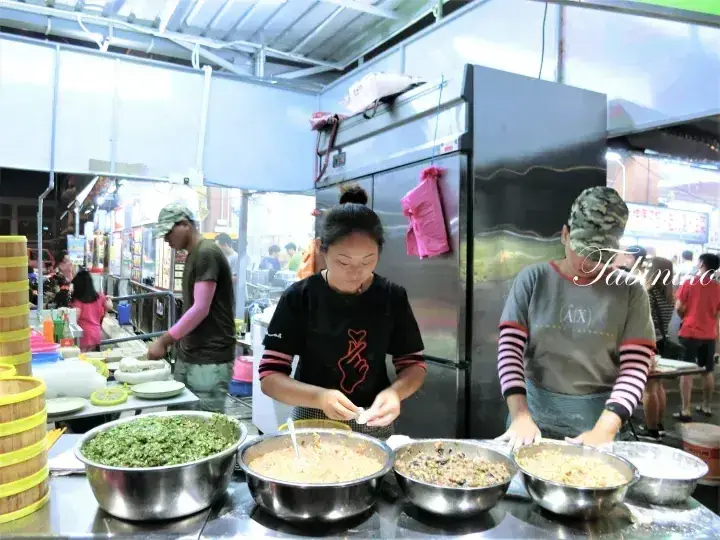 Vendors preparing traditional food at a bustling Malaysian night market stall with large bowls of ingredients.