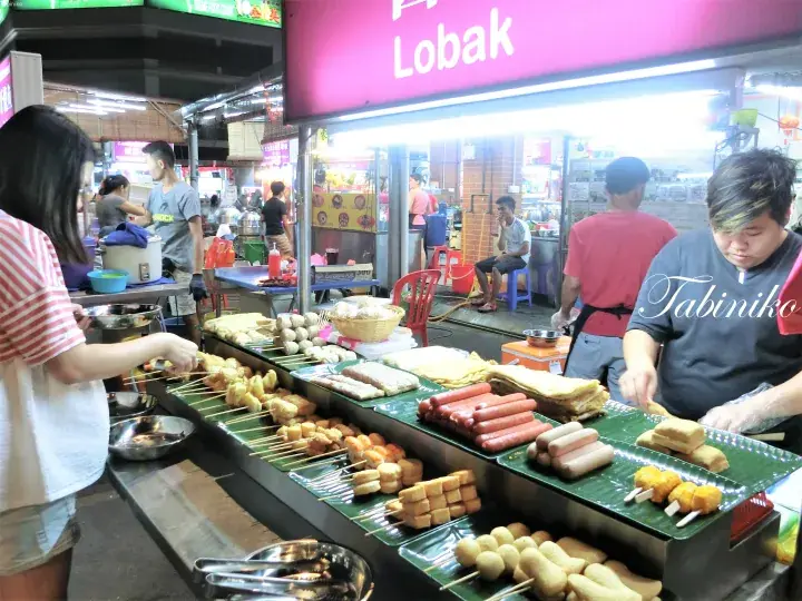 「Lobak」と書かれた看板の下で、串焼きやソーセージが並ぶ屋台の風景。