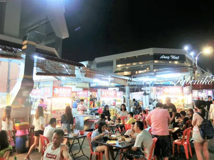 夜間の活気ある屋外フードコートで、明るい照明の下で人々がテーブル席で食事を楽しんでいる様子。