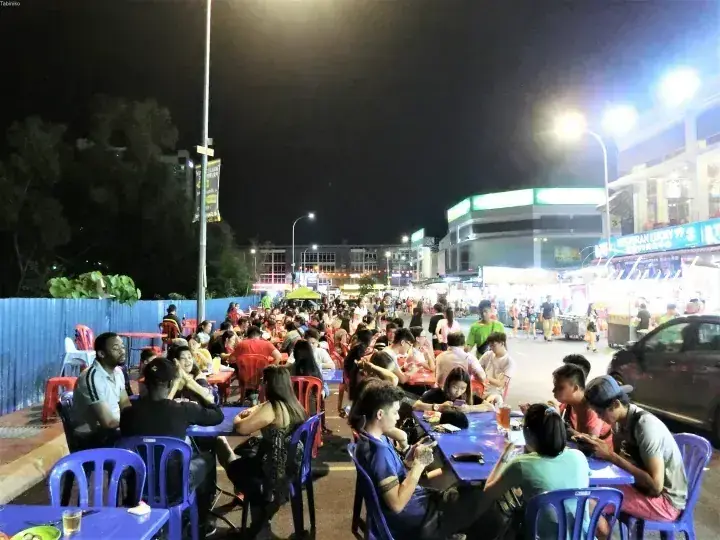 Crowd dining at night at a bustling outdoor food court with blue tables and chairs under bright lights.