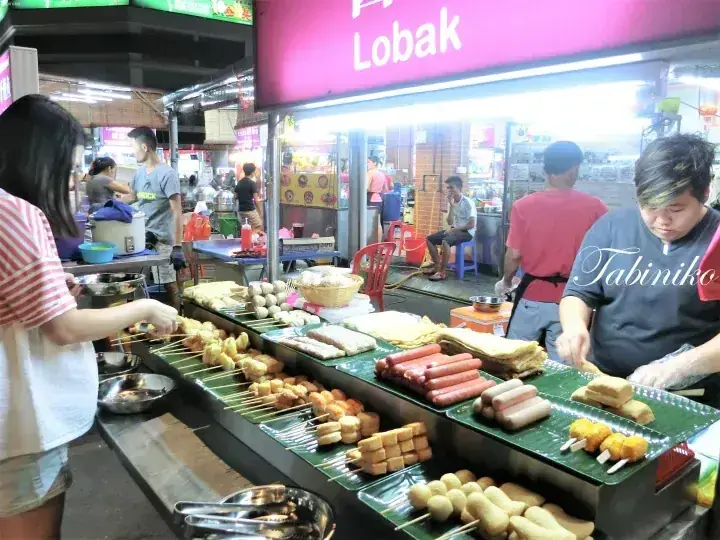 A street food stall displays various grilled skewers and sausages under a pink sign reading Lobak.