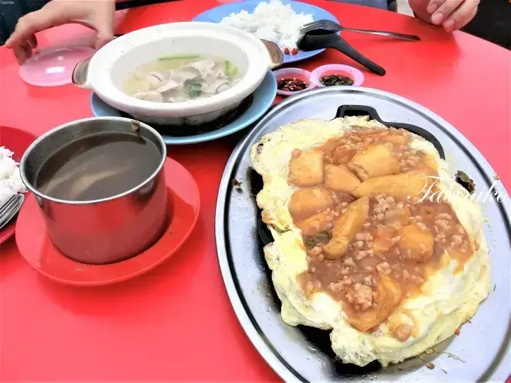 A Malaysian meal featuring a sizzling omelette with minced meat and potatoes, soup, and rice on a red table.