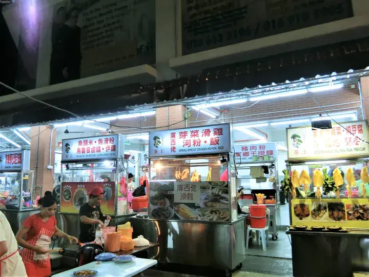 明るい照明の下で鶏飯や麺類を販売する屋台が並ぶマレーシアのハーカーセンターの夜景。
