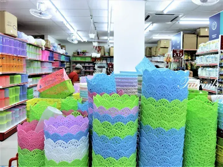 Colorful plastic baskets and storage containers stacked in a well-lit Malaysian household goods store aisle.