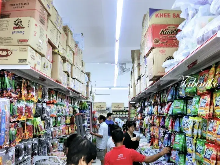 Shoppers browse aisles packed with colorful snacks and boxes of Khee San and Kuaci products in a Malaysian grocery store.