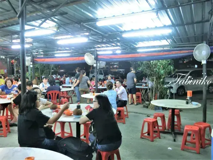 Diners enjoy a meal at a bustling open-air hawker centre at night, featuring red plastic stools and corrugated metal roofing.