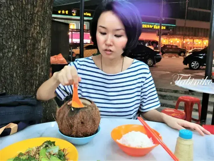 Woman eating from a coconut at Shaw Parade outdoor hawker centre in Malaysia.