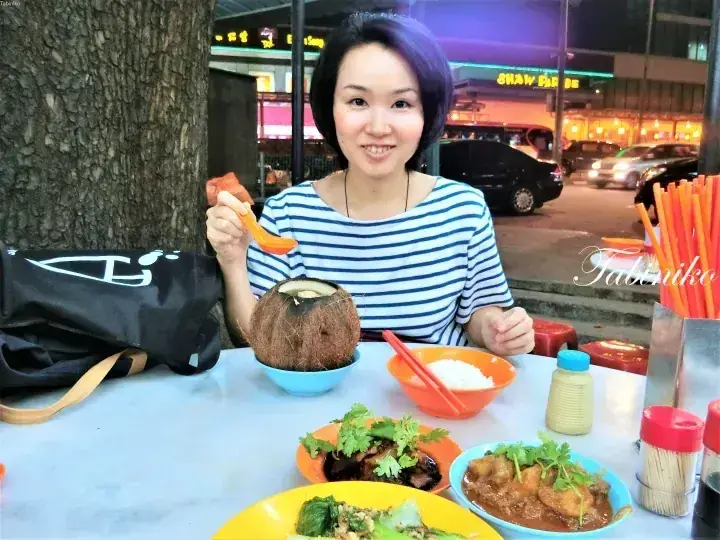 Woman with purple hair enjoying a coconut and Malaysian dishes at an outdoor hawker centre.