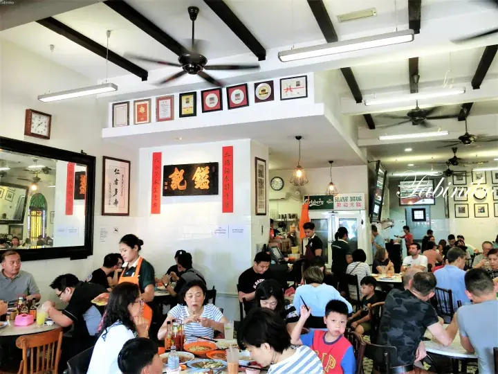 Busy dining hall at Kee Heng Kee restaurant with patrons eating at tables under ceiling fans and framed certificates.